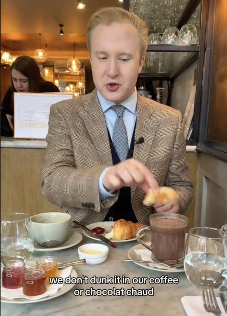British etiquette expert William Hanson sitting at a table with a croissant, coffee, hot chocolate etc.
